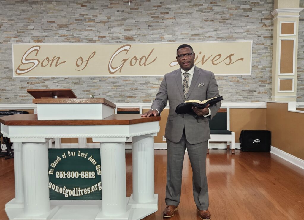 Pastor stands behind a wooden pulpit in a church, holding a Bible, with "Son of God Lives" displayed in the background. A welcoming worship setting.