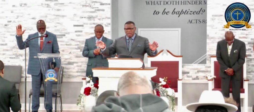 Clergy members pray during a church service, emphasizing spiritual commitment. A backdrop displays a baptism theme, highlighting the service's purpose.