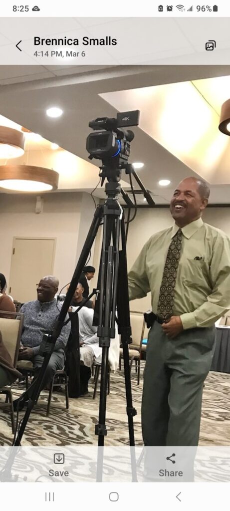 A man in a green shirt smiles while operating a camera on a tripod, capturing laughter and conversation at an event in a banquet hall.