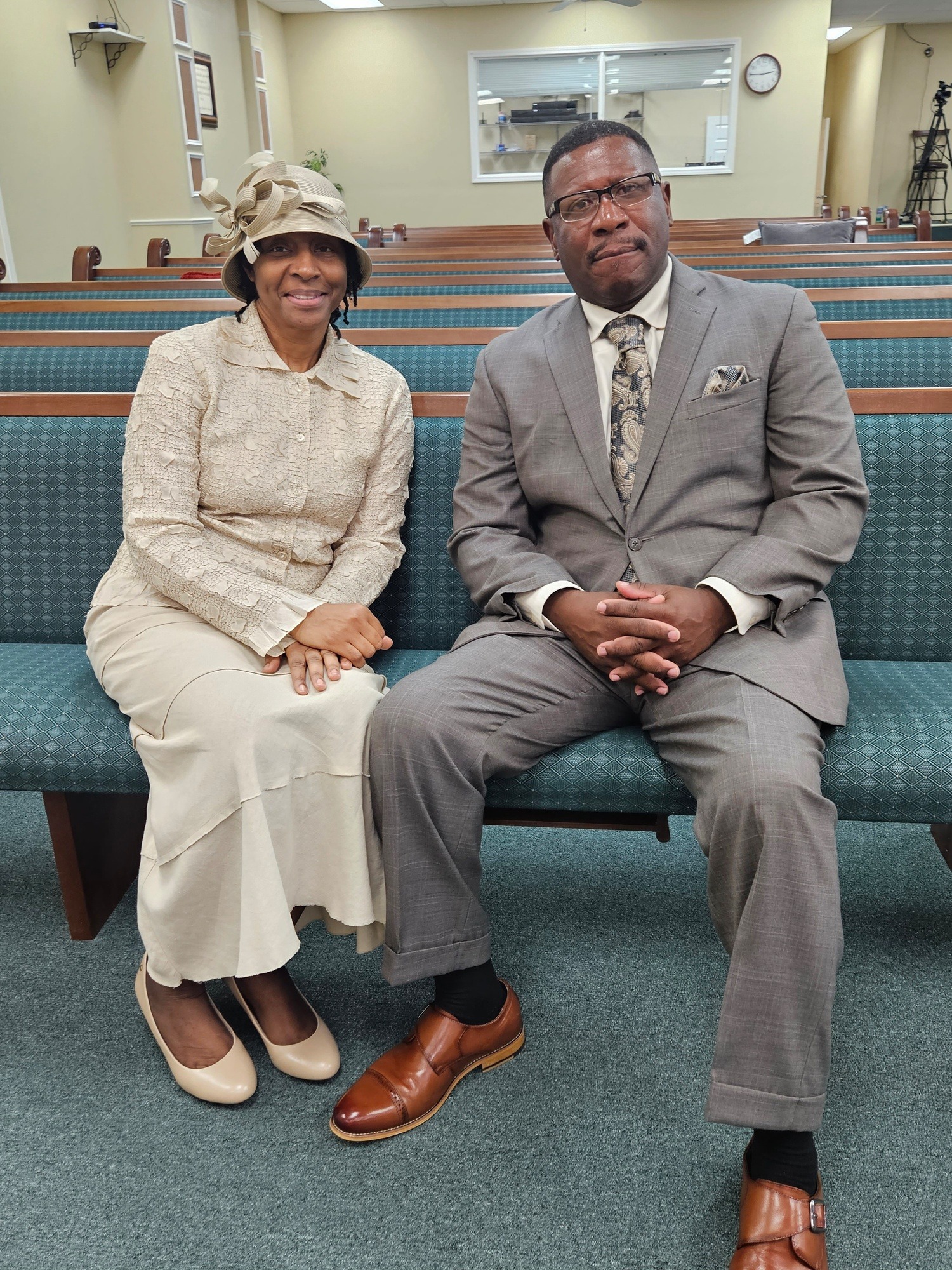 A woman in a stylish hat and beige outfit sits beside a man in a gray suit with brown shoes, both smiling in a church setting.