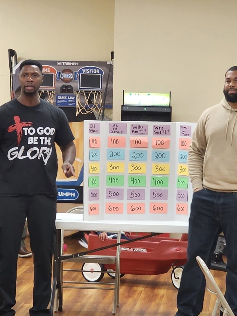 Two men stand beside a quiz board with categories related to biblical knowledge. The scene appears to be in a community center, suggesting a group activity or game.