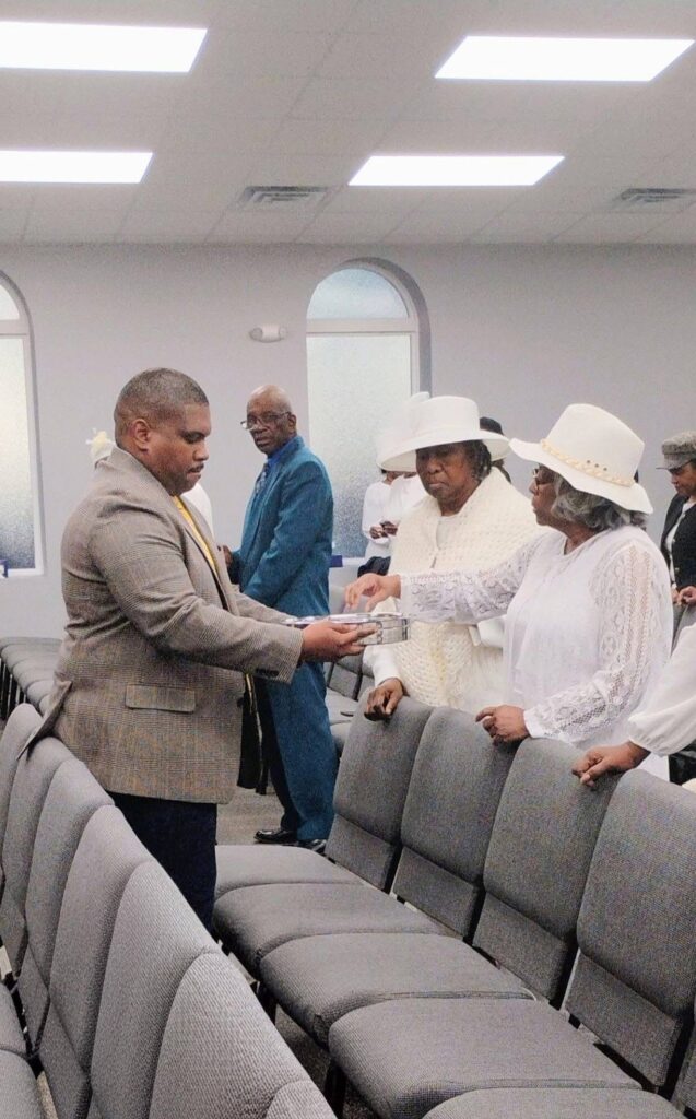 A man in a suit hands a silver tray to two women in white attire during a church gathering, highlighting a moment of fellowship and sharing.