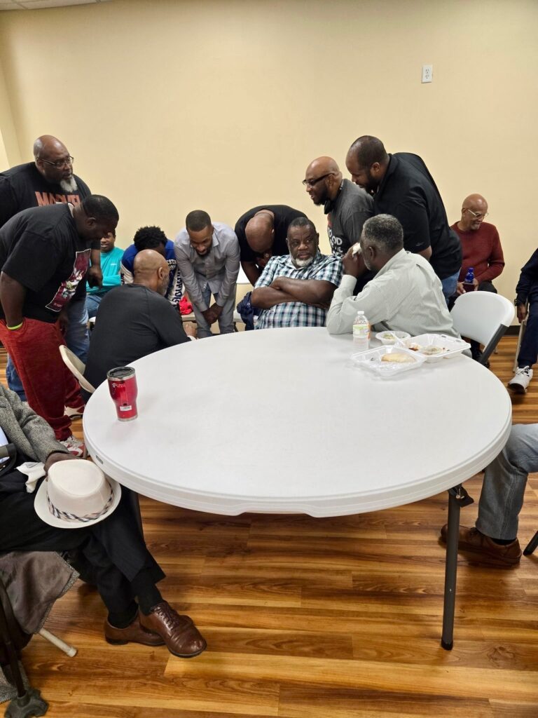 Group of men gathered around a table, engaged in conversation, with some laughing and others listening attentively. A casual, friendly atmosphere.
