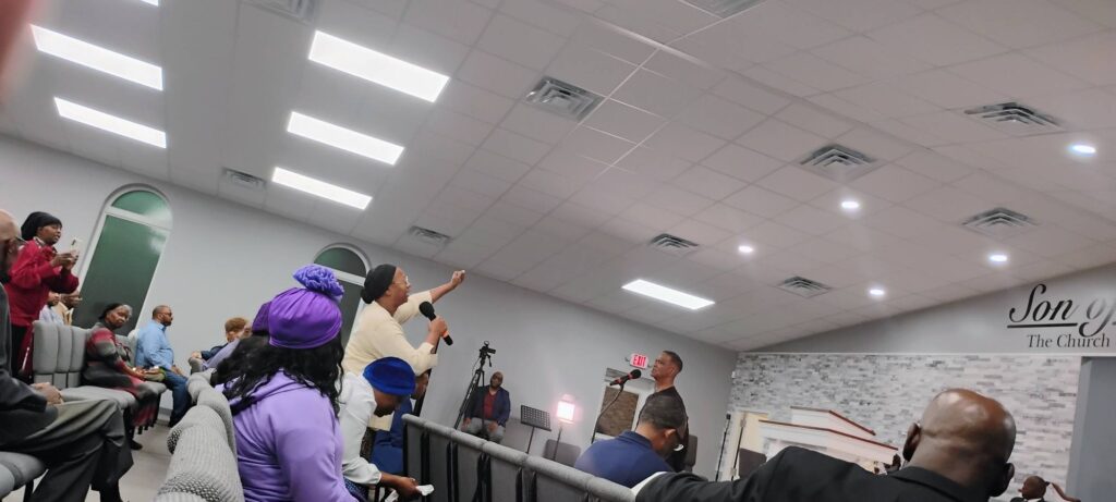A church gathering with attendees engaged in conversation; a woman, microphone in hand, speaks passionately to a man at the front.