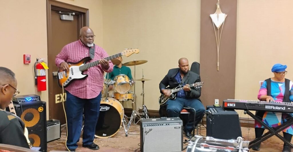 A live band performs indoors, featuring a bassist in a red checkered shirt, a drummer, a guitarist in a blazer, and a woman playing a keyboard.