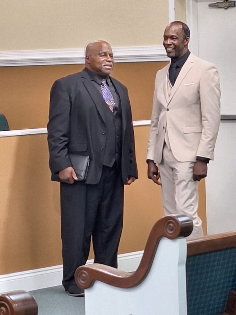 Two men are smiling and conversing in a formal setting. One is dressed in a black suit, while the other wears a light beige suit, suggesting a celebratory or professional occasion.