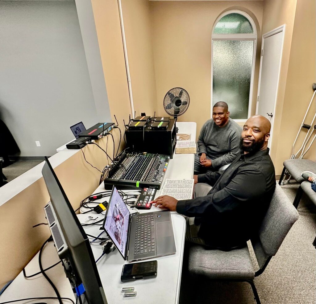 Two men work at a sound and video control station, adjusting equipment and monitoring screens. The setting appears to be a church or community event.