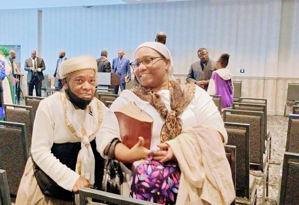 An elderly man and a woman smile together in a conference setting, both dressed in formal attire and with items like a book and a coat.