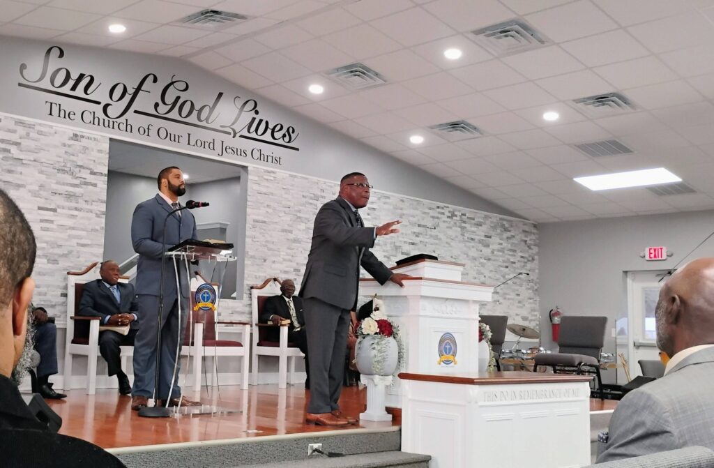 Two men speaking at a church, one delivering a sermon while the other listens. The setting is modern with religious symbols and seating for the congregation.