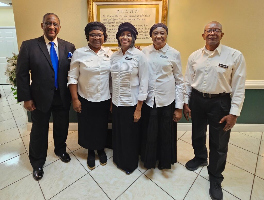 A group of five individuals poses together, dressed in formal attire, smiling warmly. They stand near a framed biblical quote, likely representing a church or community event.