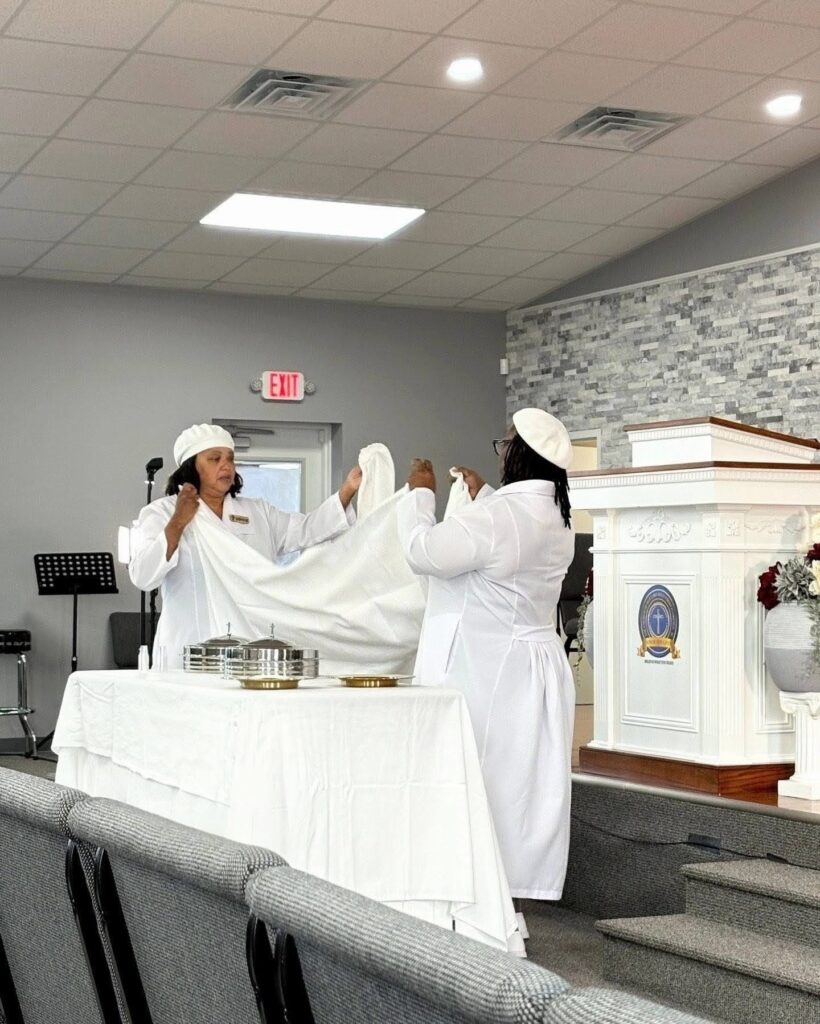 Two women in white attire prepare a table with silver communion trays in a church setting, highlighting a moment of religious observance.