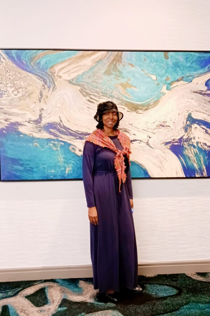 Woman in a long purple dress and a black hat stands smiling against a colorful abstract painting, showcasing her stylish outfit and confidence.
