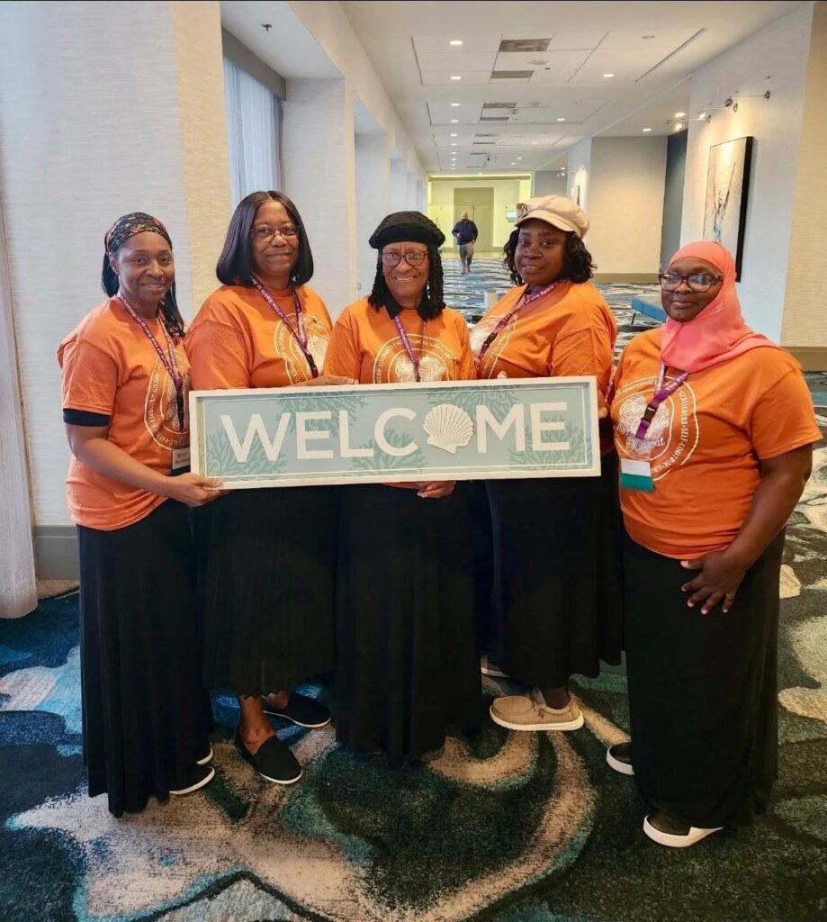 Five women in orange t-shirts stand together holding a sign that reads "WELCOME," set in a well-lit conference space, promoting a welcoming atmosphere.