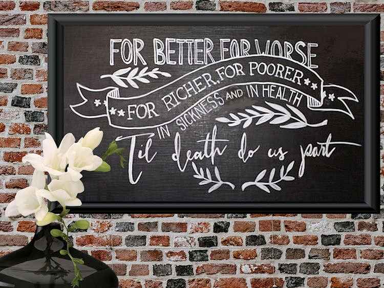 Chalkboard art with white cursive text reading wedding vows, adorned with floral accents. A vase with white flowers sits in front of a brick wall.