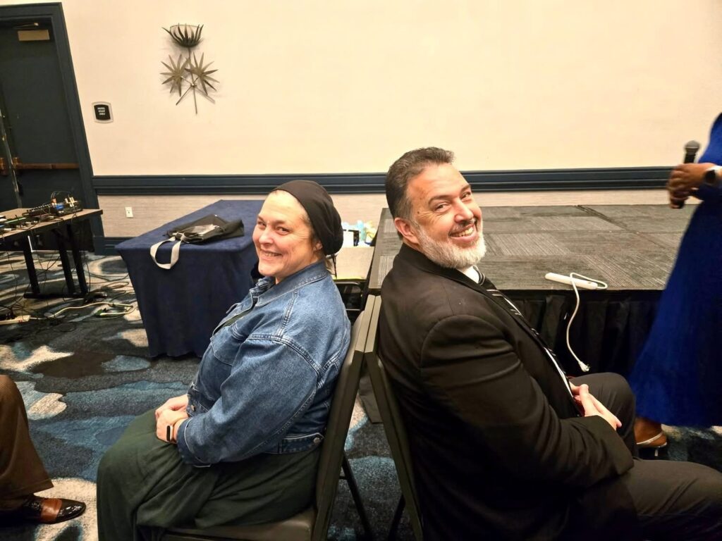 Two smiling individuals sit back-to-back on chairs in a conference setting, surrounded by event equipment and a stage backdrop.