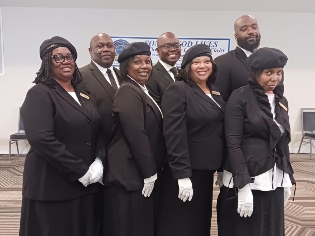 A group of nine individuals dressed in formal black attire with white gloves, posing together, likely for a church or community event.