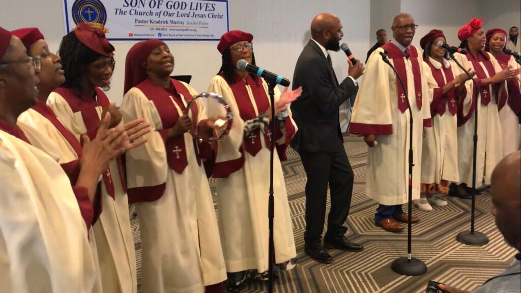 A gospel choir performs joyfully, dressed in white and maroon robes, as they sing into microphones at a church event.