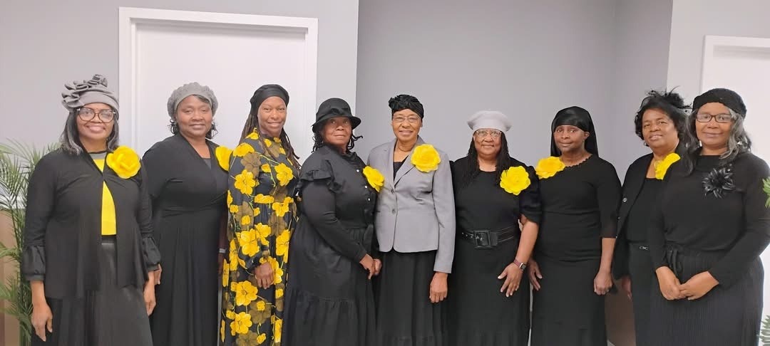 A group of ten women dressed in black with yellow accents, standing together in a bright room, showcasing unity and elegance during a special event.