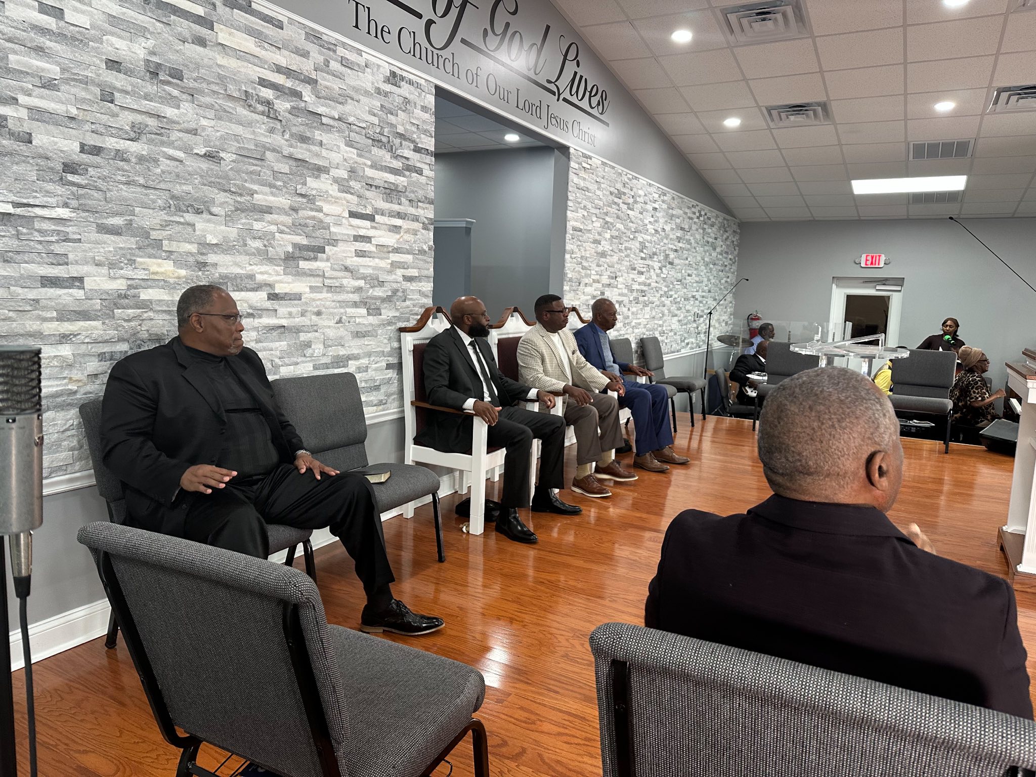 A group of men in suits seated in a church setting, facing an unseen speaker. The interior features modern decor with a gray stone wall, enhancing the spiritual atmosphere.