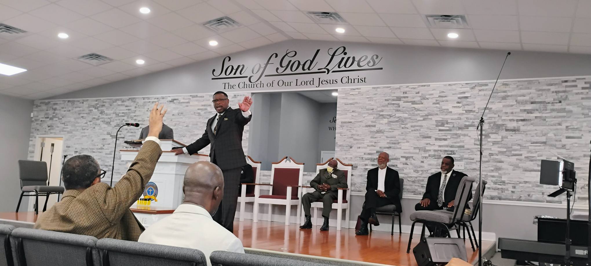 A pastor gestures while speaking to the congregation in a modern church setting, with attentive attendees seated behind him.
