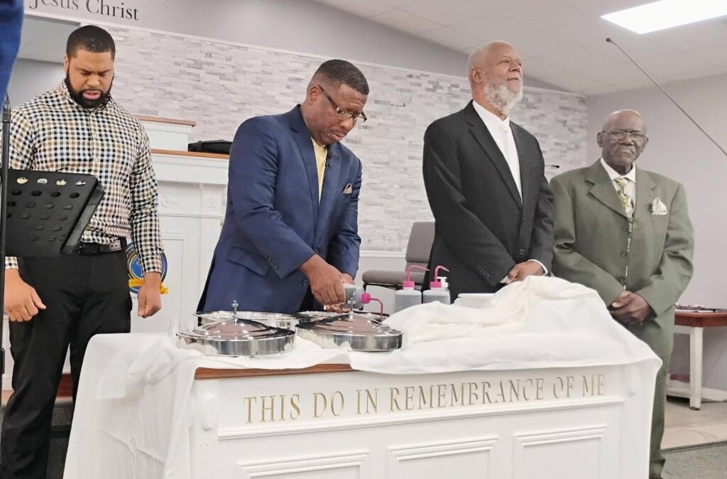 Four men stand reverently at a church altar during a communion service, as one prepares the communion elements. The text on the altar reads, "THIS DO IN REMEMBRANCE OF ME."