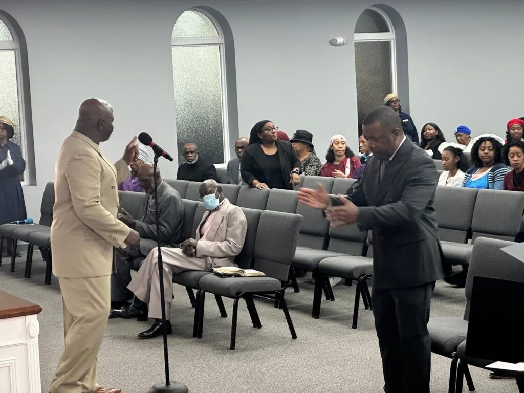 A man in a beige suit speaks passionately into a microphone while another man gestures animatedly. The audience, dressed in formal attire, listens attentively in a church setting.