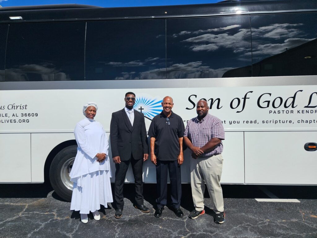 Four individuals, dressed in formal attire, stand in front of a bus with "Son of God Life" logo, highlighting community engagement and outreach.