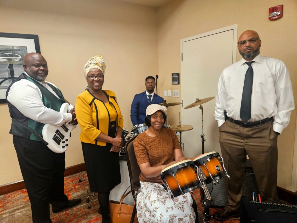 A musical group poses together in a professional setting, featuring a bassist, percussionist, and others, showcasing their instruments and attire, ready to perform.