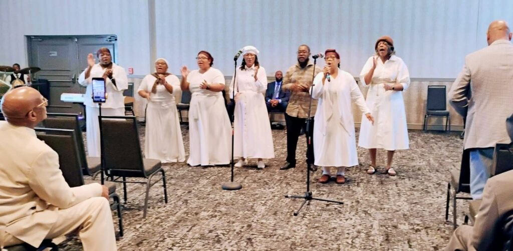A diverse group of singers performs lively gospel music in white attire, engaging the audience in a joyful worship setting.
