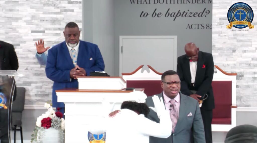 Clergy members gather in a church setting, praying for a participant in baptism, symbolizing faith and community support.