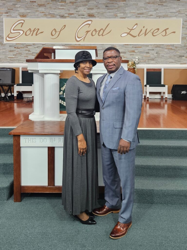 A woman in a gray dress and hat stands beside a man in a gray suit at a church podium, showcasing a welcoming worship atmosphere.