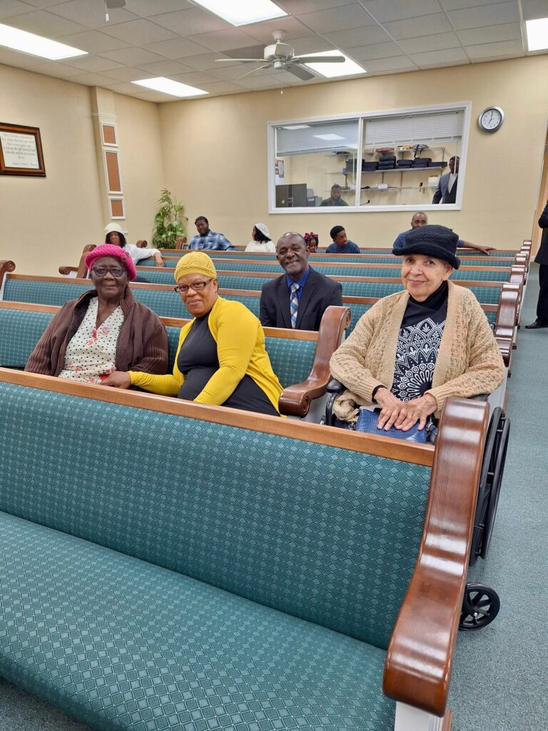 Four individuals, including three women with colorful hats, sit in a church-like setting, engaged in conversation. The image highlights community and togetherness.