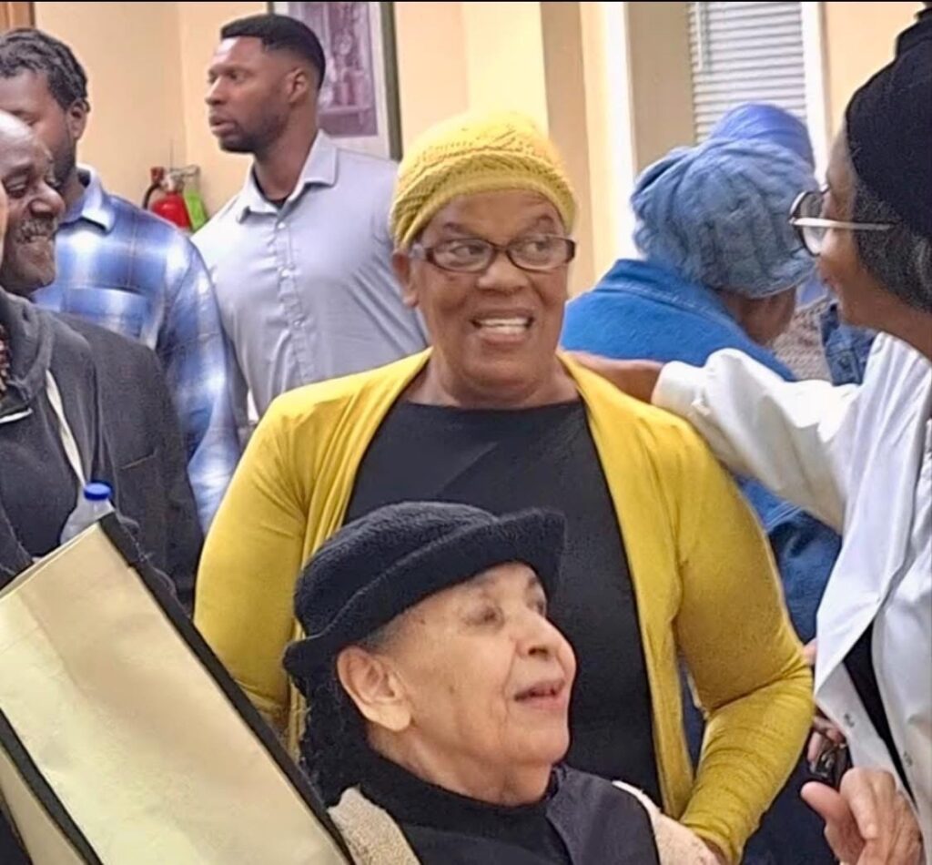 A smiling group of people, including a woman in a yellow sweater and a black hat, interact warmly, highlighting community connection and joy.