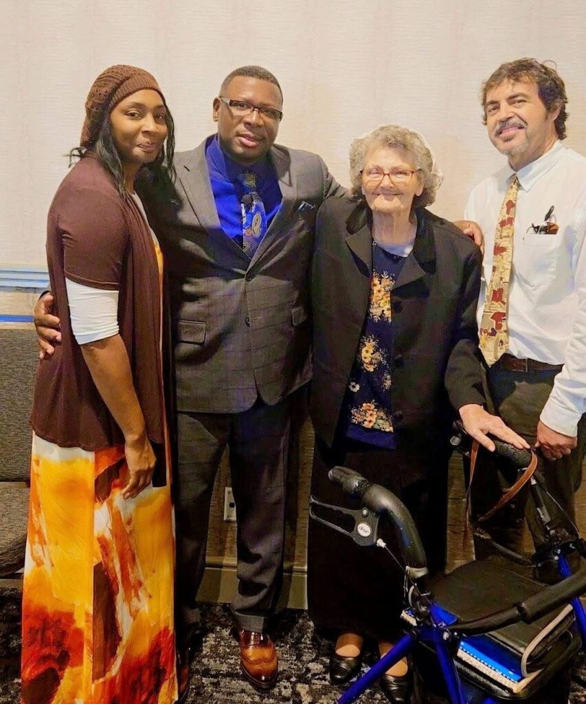 A group of four people poses together, smiling. They are dressed in formal attire, with a walker visible beside an elderly woman in a black jacket, suggesting a family event or celebration.