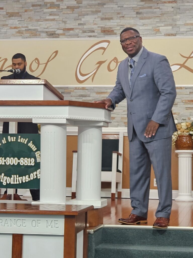 A pastor in a gray suit stands next to a podium in a church, smiling while addressing the congregation. A microphone and another speaker are nearby.