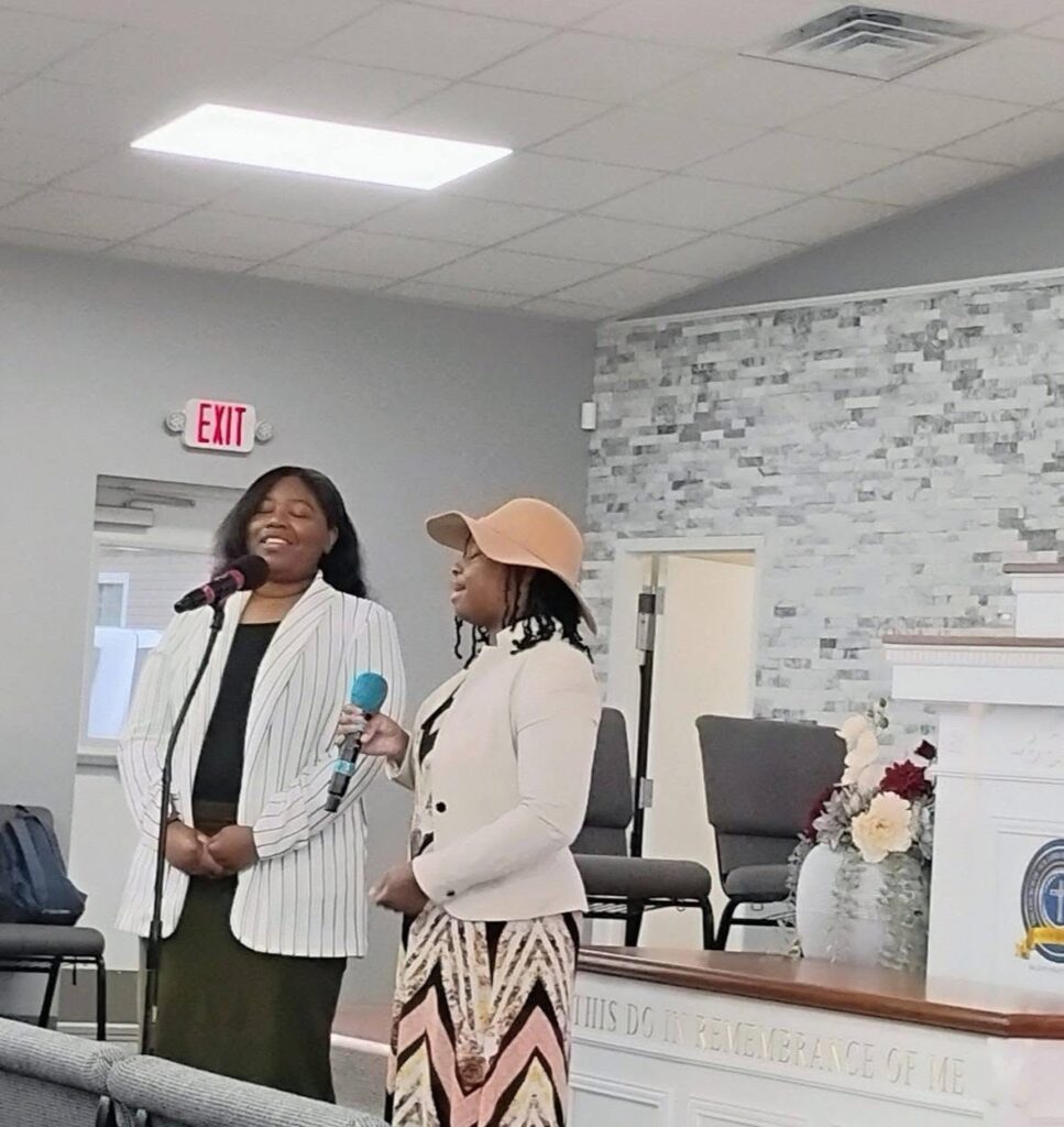 Two women sing at a church event, one holding a blue microphone. The background features a modern interior with decorative elements.