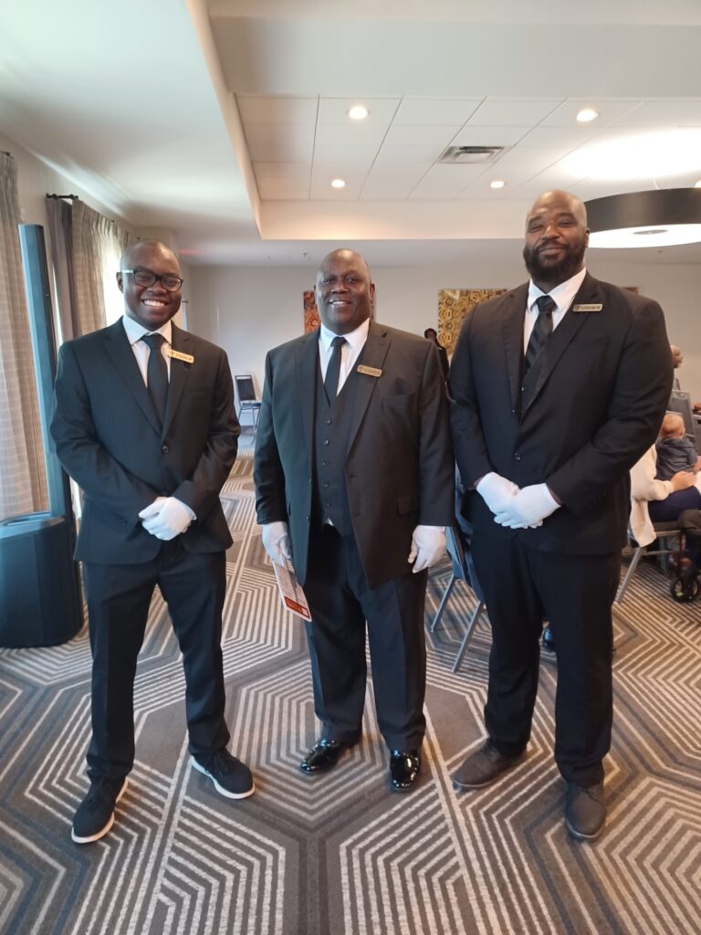 Three formal staff members in black suits and white gloves stand in a well-lit event space, ready to assist guests at a gathering.