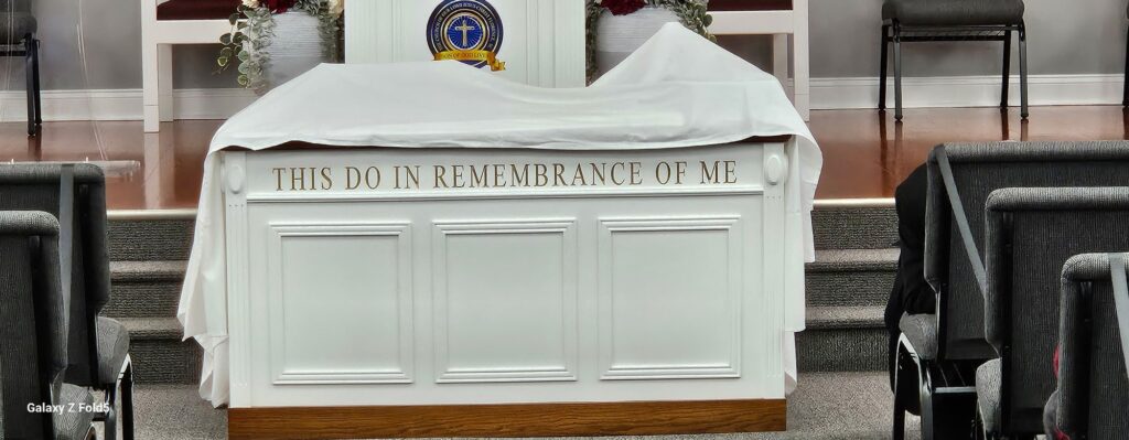 Altar with a white cloth and the inscription "THIS DO IN REMEMBRANCE OF ME," set in a church interior, symbolizing communion and remembrance.