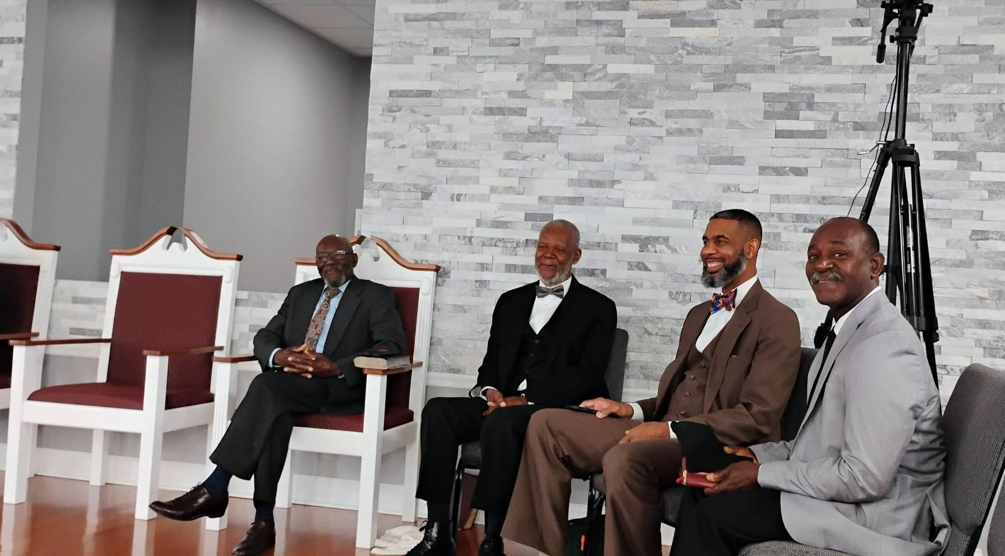 Four men in formal attire sit together, smiling, in a modern, light-filled room with a stone wall backdrop. The image captures a moment of camaraderie.