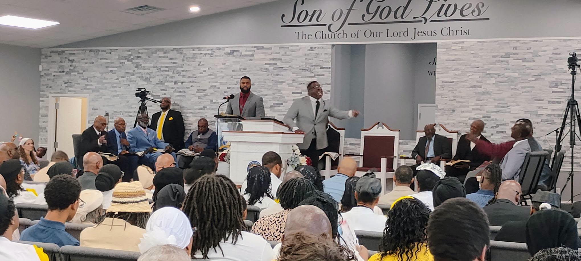 A lively church service at Son of God Lives features a speaker engaging the audience, with a diverse congregation attentively listening.