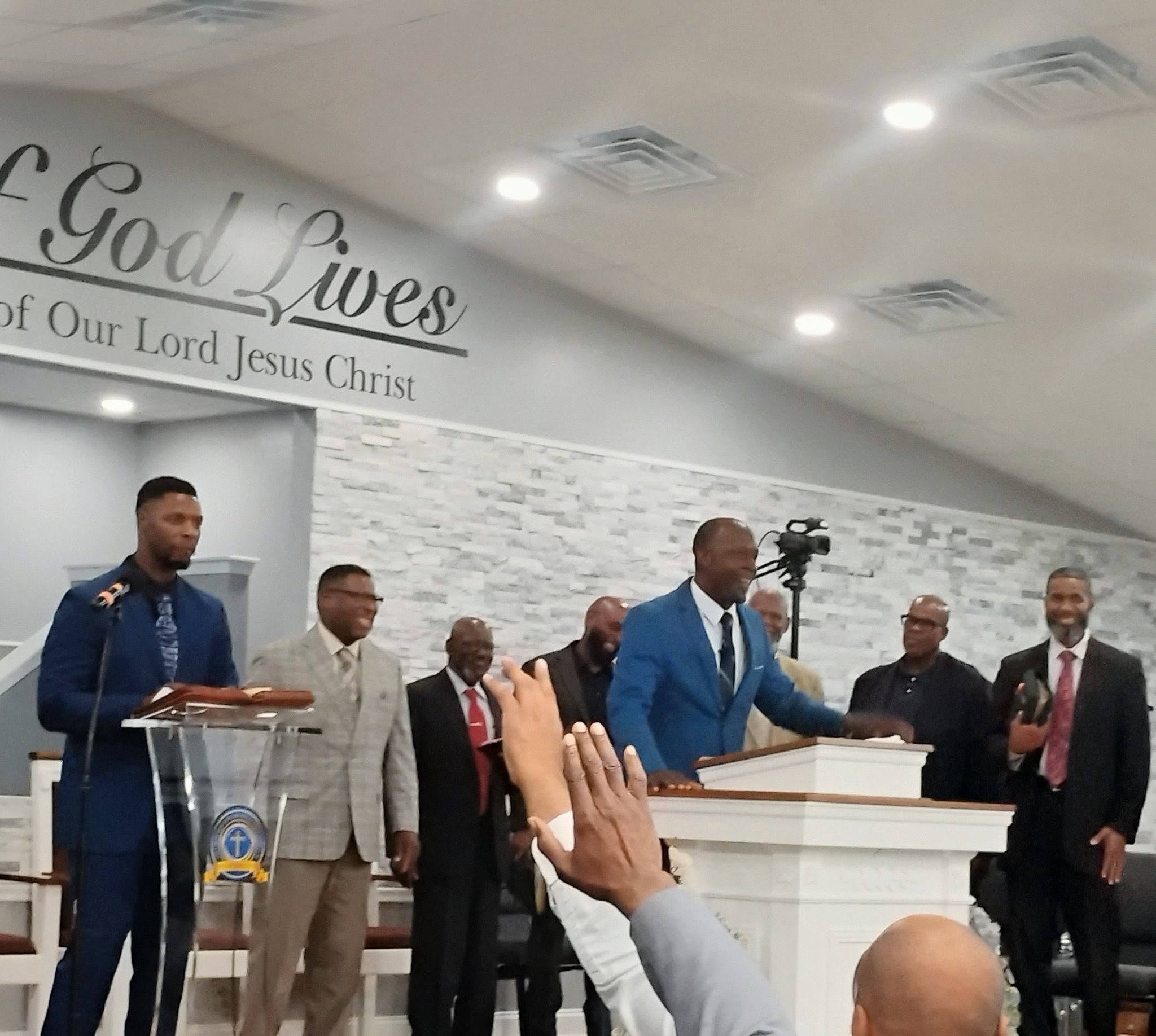 Pastors and congregation members gather in a church during a service, with one pastor preaching passionately at a podium.