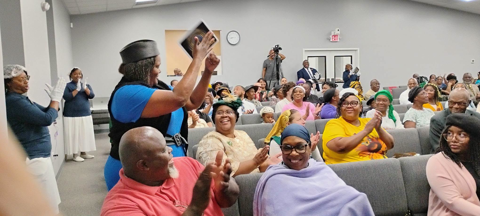 A lively community event featuring a diverse audience clapping and celebrating, with a woman in a blue shirt joyously raising her hands in the foreground.