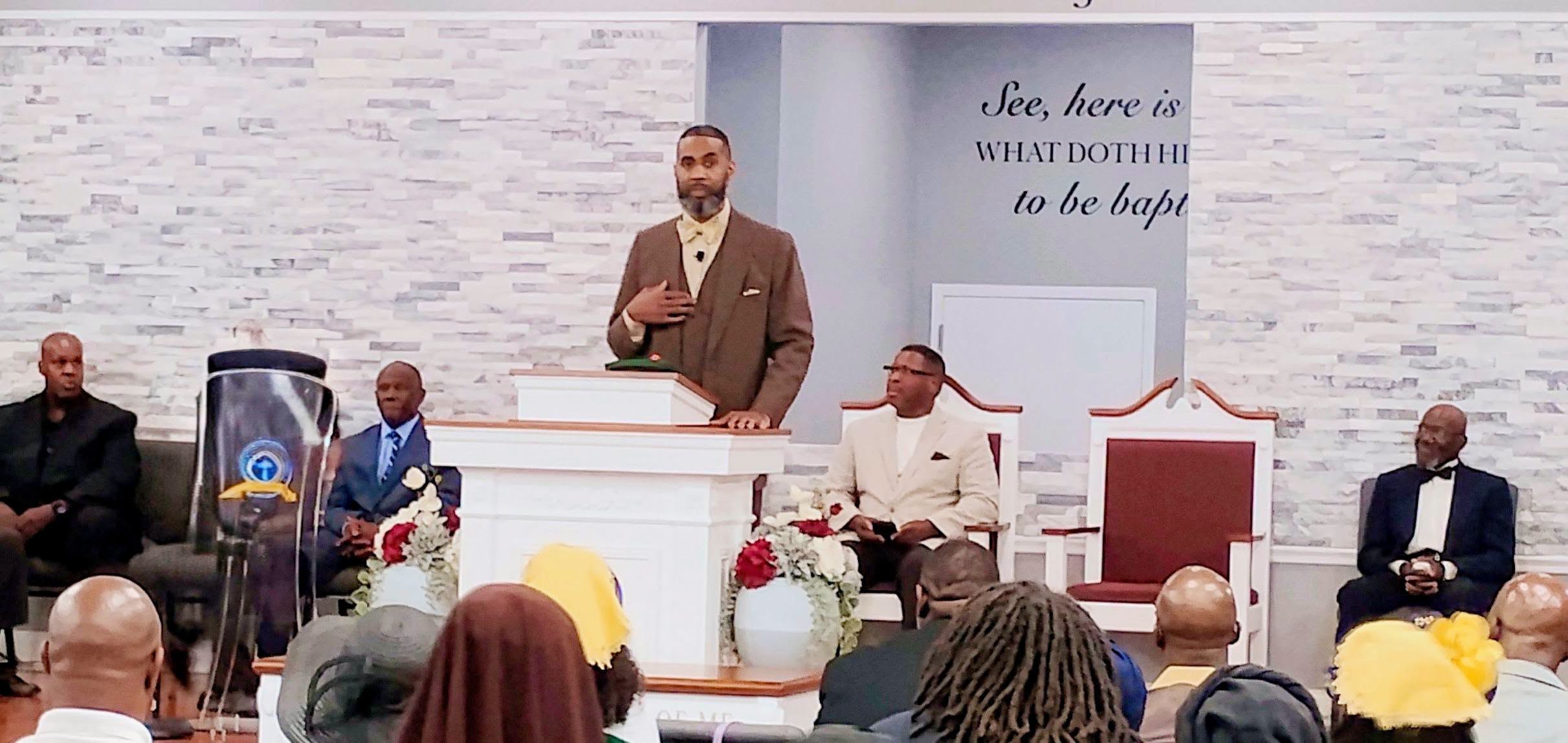 A pastor speaks at a church service, addressing a diverse congregation. Floral arrangements decorate the podium, enhancing the spiritual atmosphere.