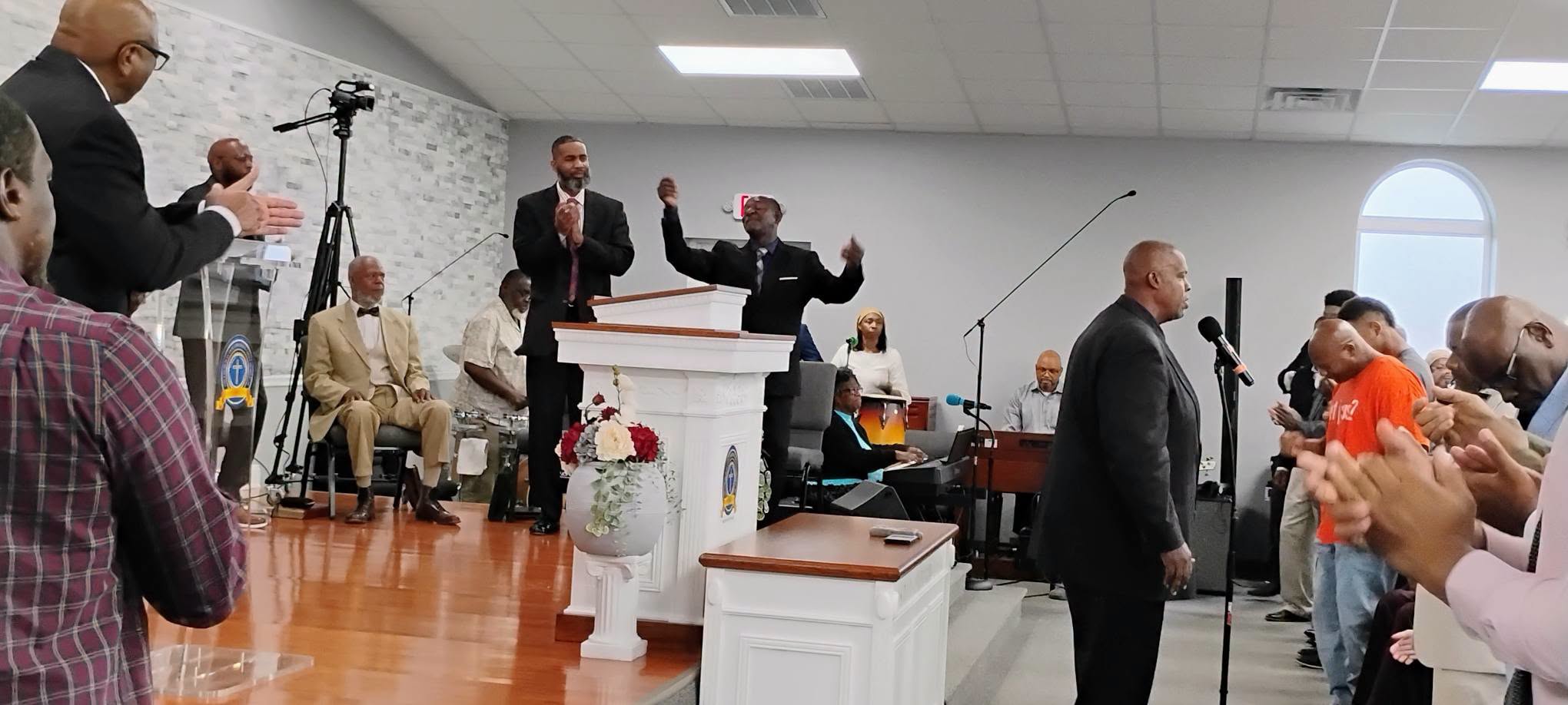 Worship service in a church, featuring multiple speakers, musicians, and an engaged congregation. The atmosphere is lively and celebratory.