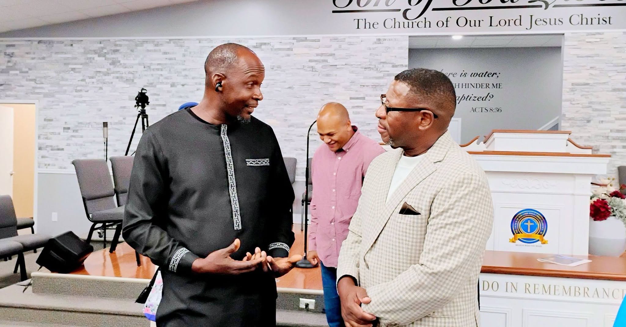 Two men engaged in conversation at a church gathering, with a third person smiling in the background. The setting features a modern church interior.