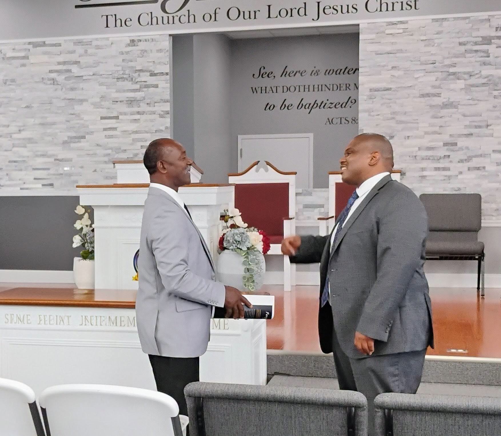Two men in business attire engage in a friendly conversation in a church setting, with a pulpit and floral arrangements in view.
