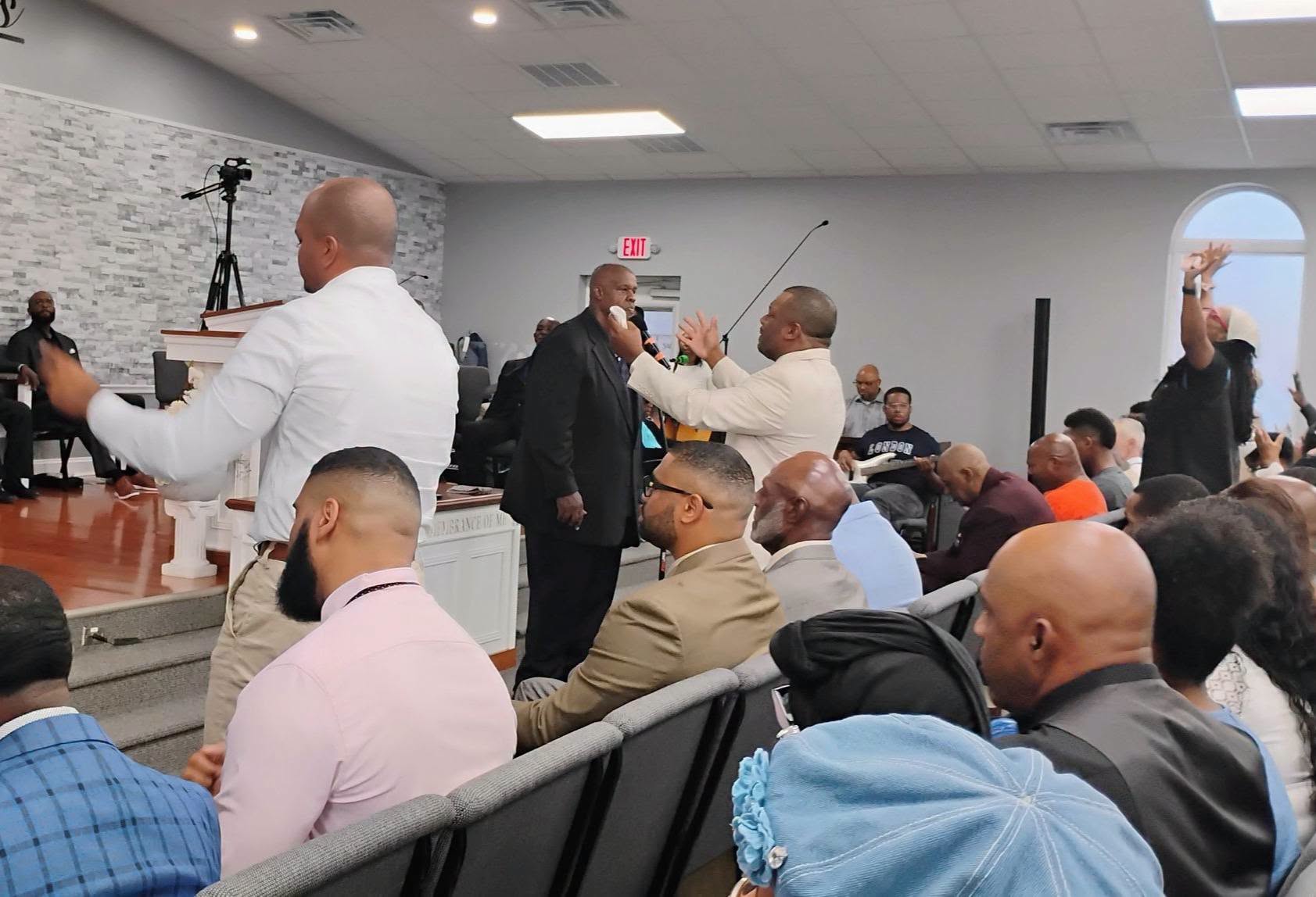 A diverse group of people engages in a lively church service. Two men stand at the front; one speaks passionately while attendees listen intently.