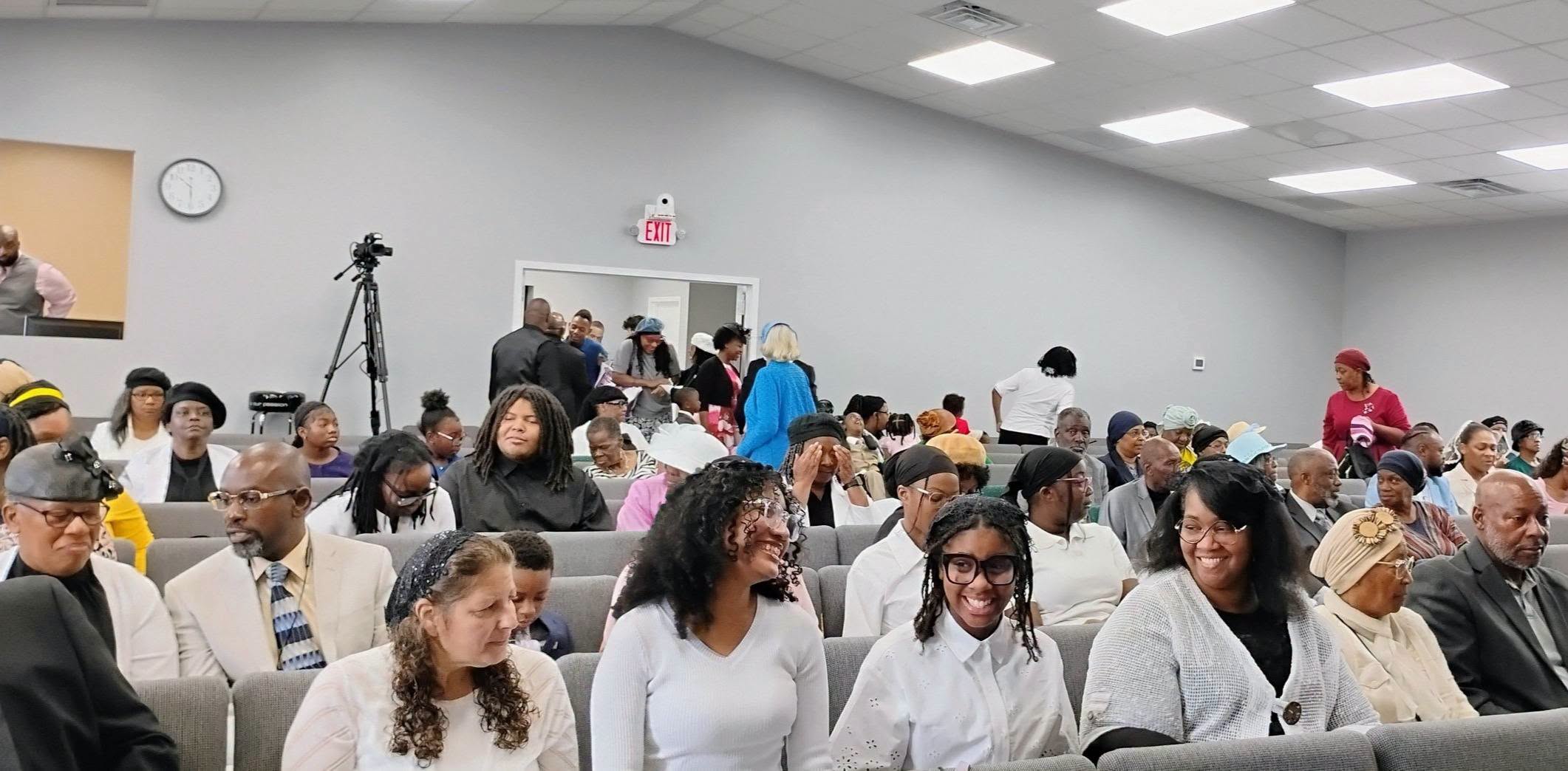 A diverse congregation of individuals seated in a well-lit hall, engaging with one another, likely in preparation for a community event or service.